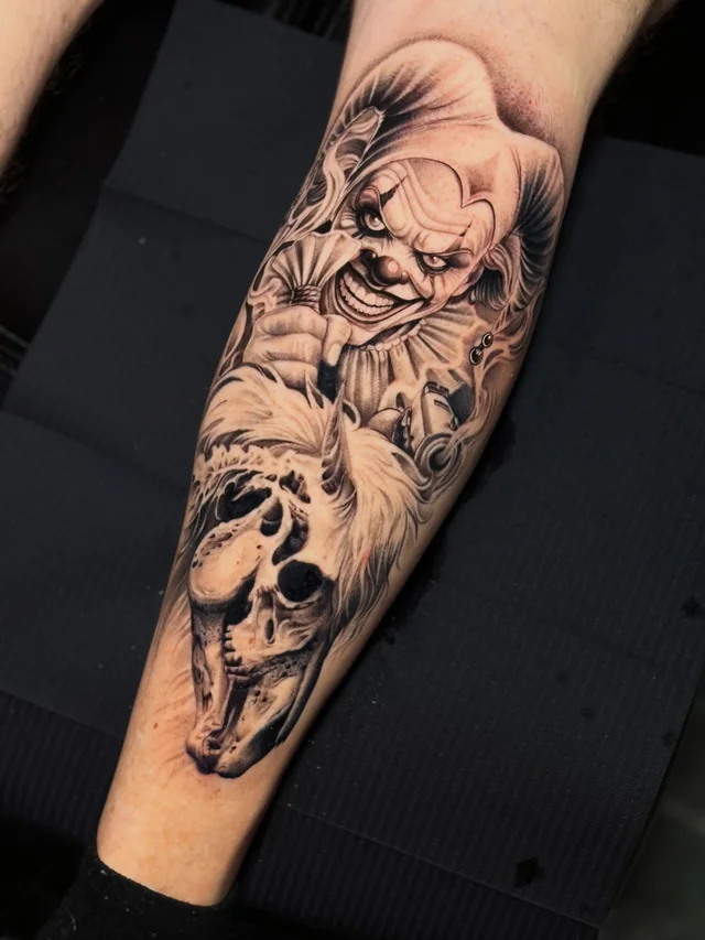 Horror-style clown portrait above an animal skull in a black and grey forearm tattoo.