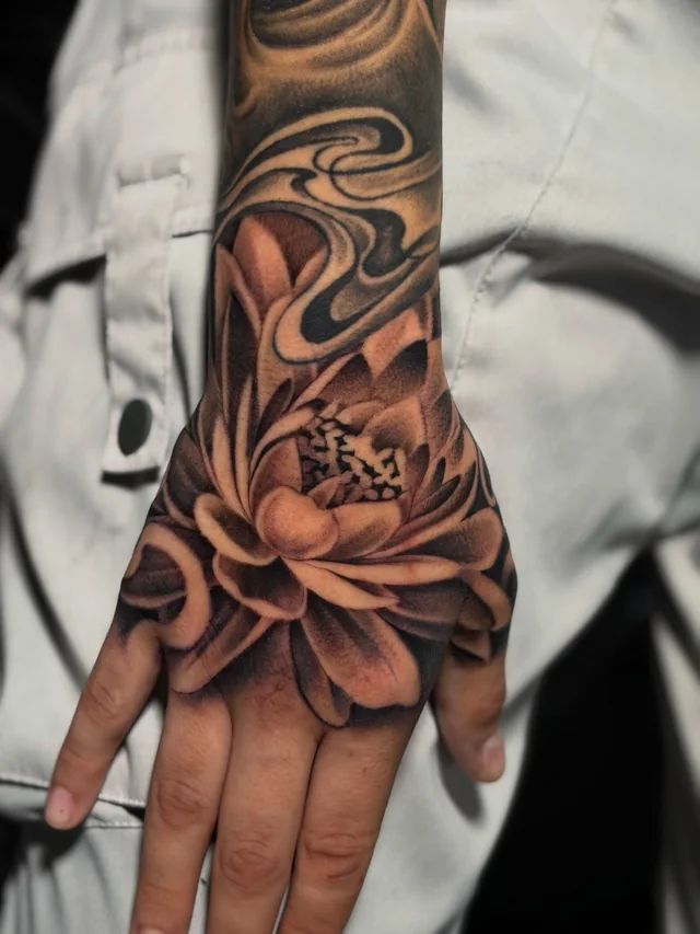 Shaded lotus-like flower tattoo on a hand beside flowing forearm tattoo lines.