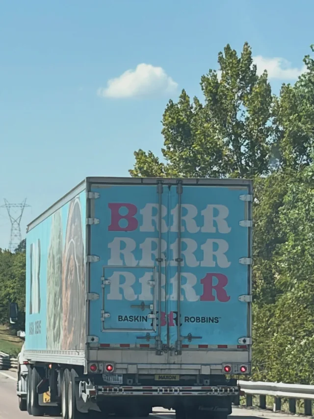 Turquoise ice-cream truck on a road beneath blue sky and trees.