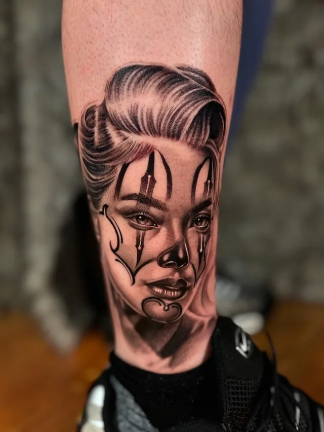Female facial portrait framed by ornamental lines in a black and grey lower-leg tattoo.