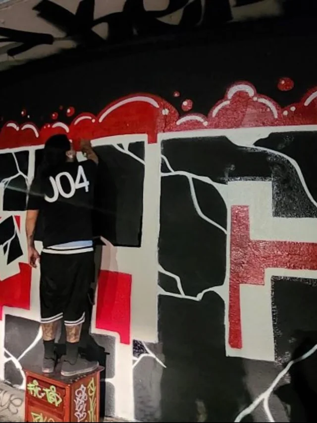 Person in a black shirt and shorts standing beside a wall of graffiti.