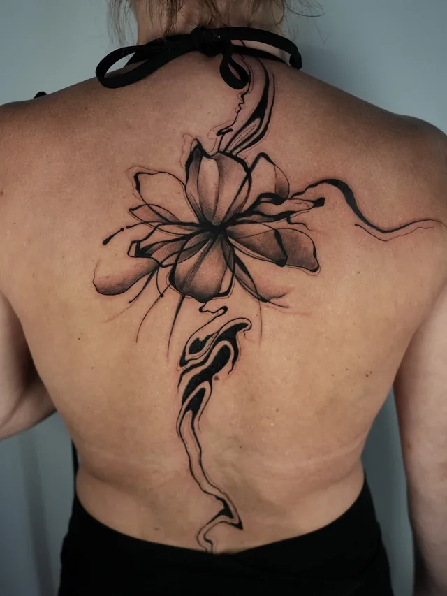 Back tattoo with a large shaded flower and flowing black ribbon-like forms extending toward the lower back.