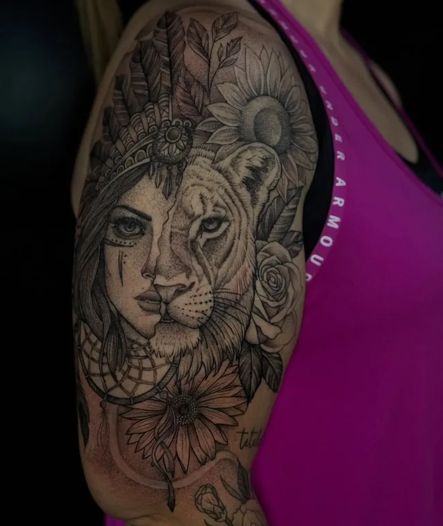Upper-arm tattoo combining half a human face and half a lion face, framed by feathers and large flowers.