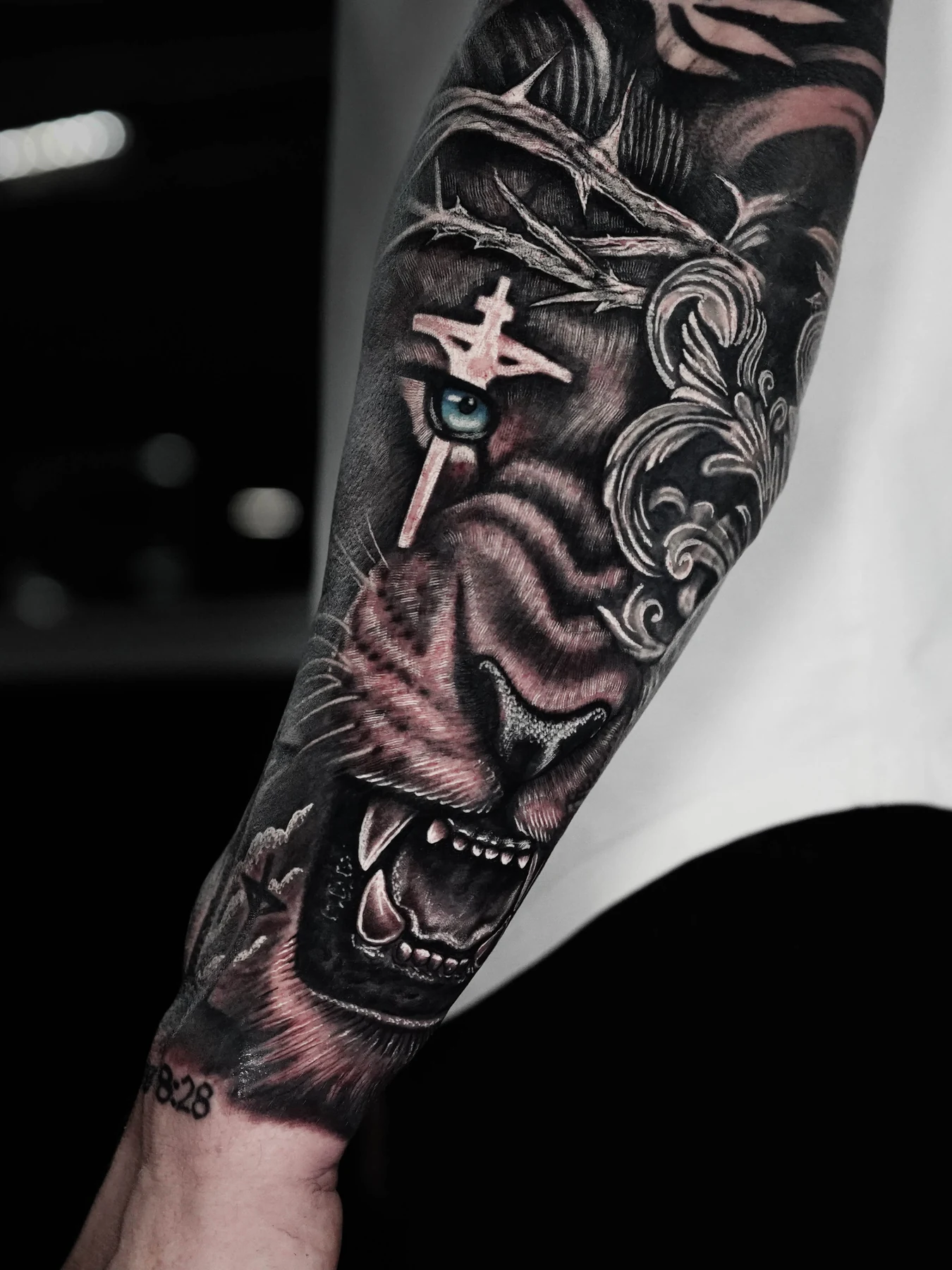 A black-and-gray forearm tattoo featuring a lion with a crown of thorns and a cross.