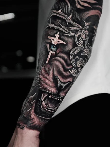 A black-and-gray forearm tattoo featuring a lion with a crown of thorns and a cross.