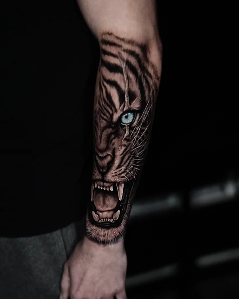 A close-up tiger face tattoo on the forearm, rendered in black and gray with a pale blue eye.