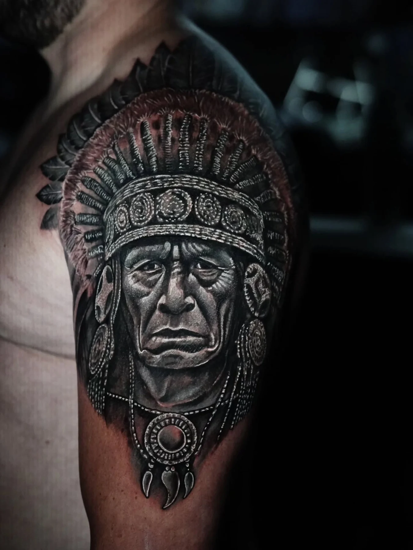 A black-and-gray upper-arm portrait of a figure wearing a detailed feathered headdress.
