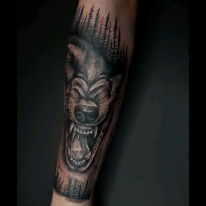 A black-and-gray wolf tattoo framed by pine-tree silhouettes.