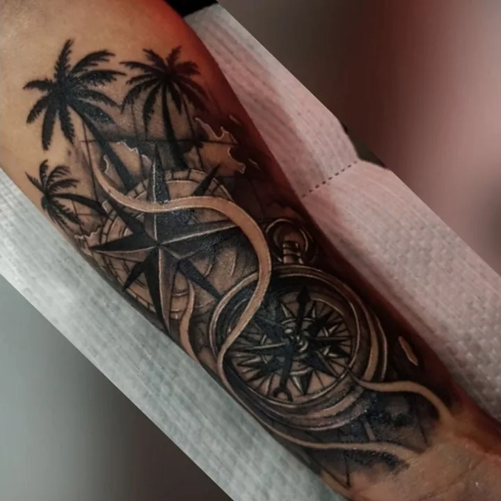 A black-and-gray compass tattoo combined with palm trees and a coastal scene.