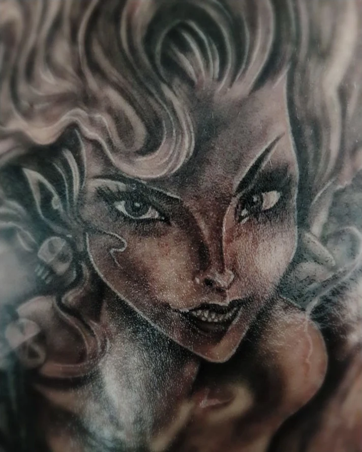 A stylized face tattoo in black and gray, surrounded by flowing hair and smoky shapes.
