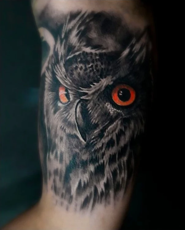 An owl portrait tattoo with vivid orange eyes set against dark, layered feathers.