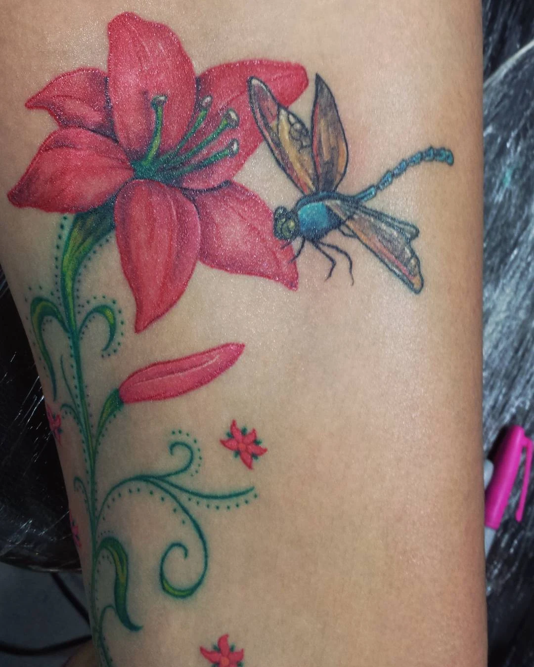 A colorful floral tattoo with a red flower, a blue-green dragonfly, and curling plant stems.