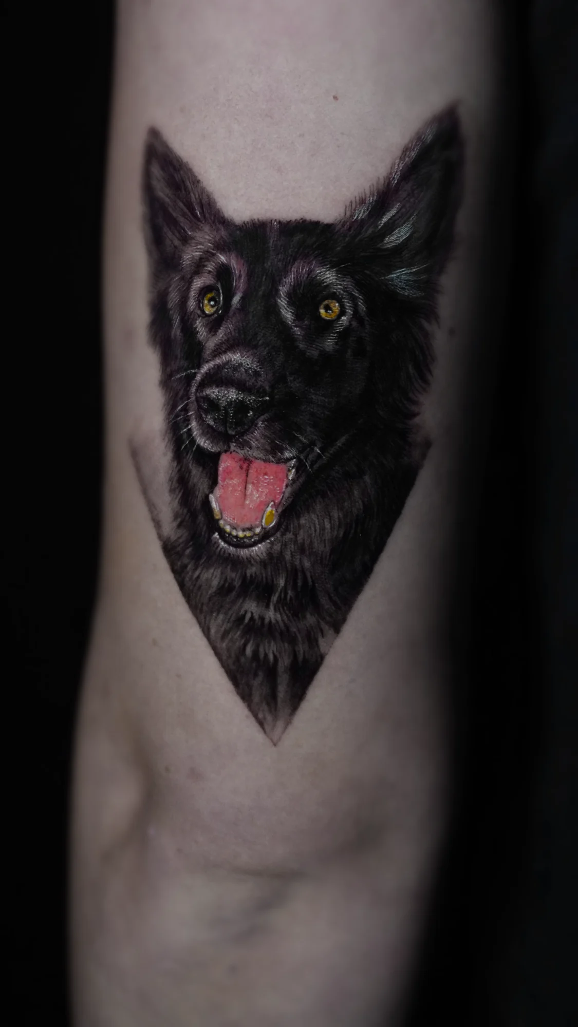 A portrait tattoo of a dark-coated dog with upright ears, bright eyes, and its tongue showing.