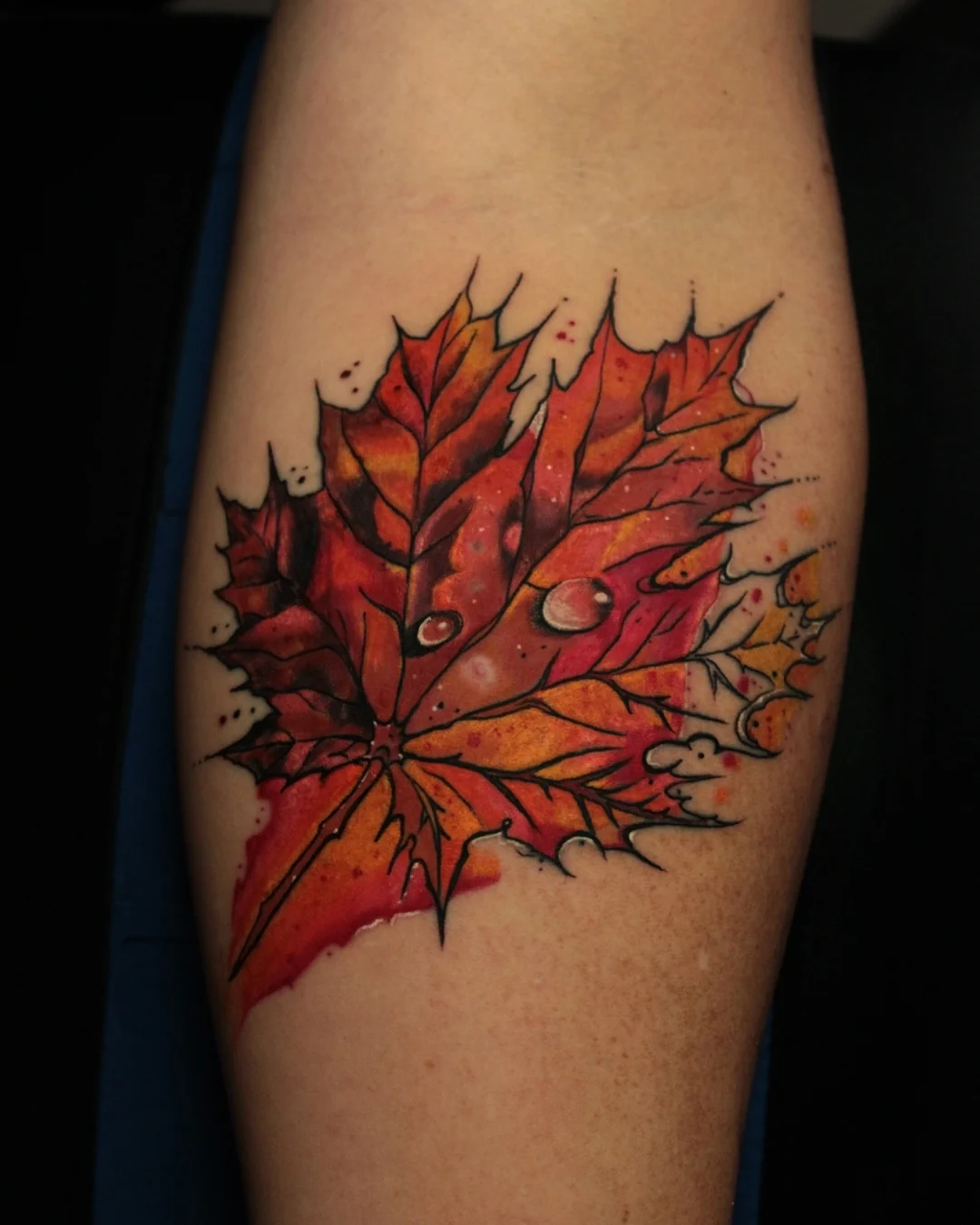 A red and orange autumn maple leaf tattoo with dark veins, pointed edges and two shaded water droplets.