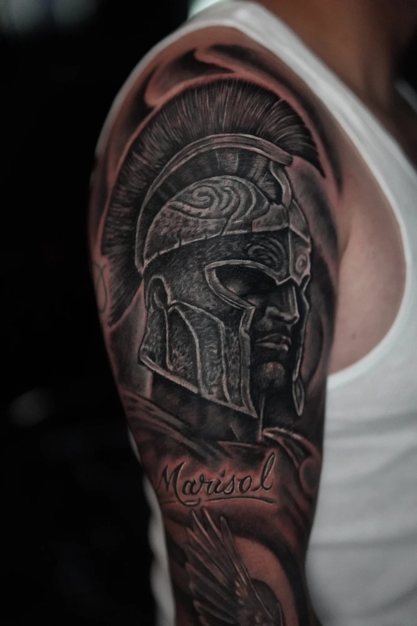 A black-and-gray upper-arm tattoo of a crested warrior helmet with engraved armor and a ribbon below.