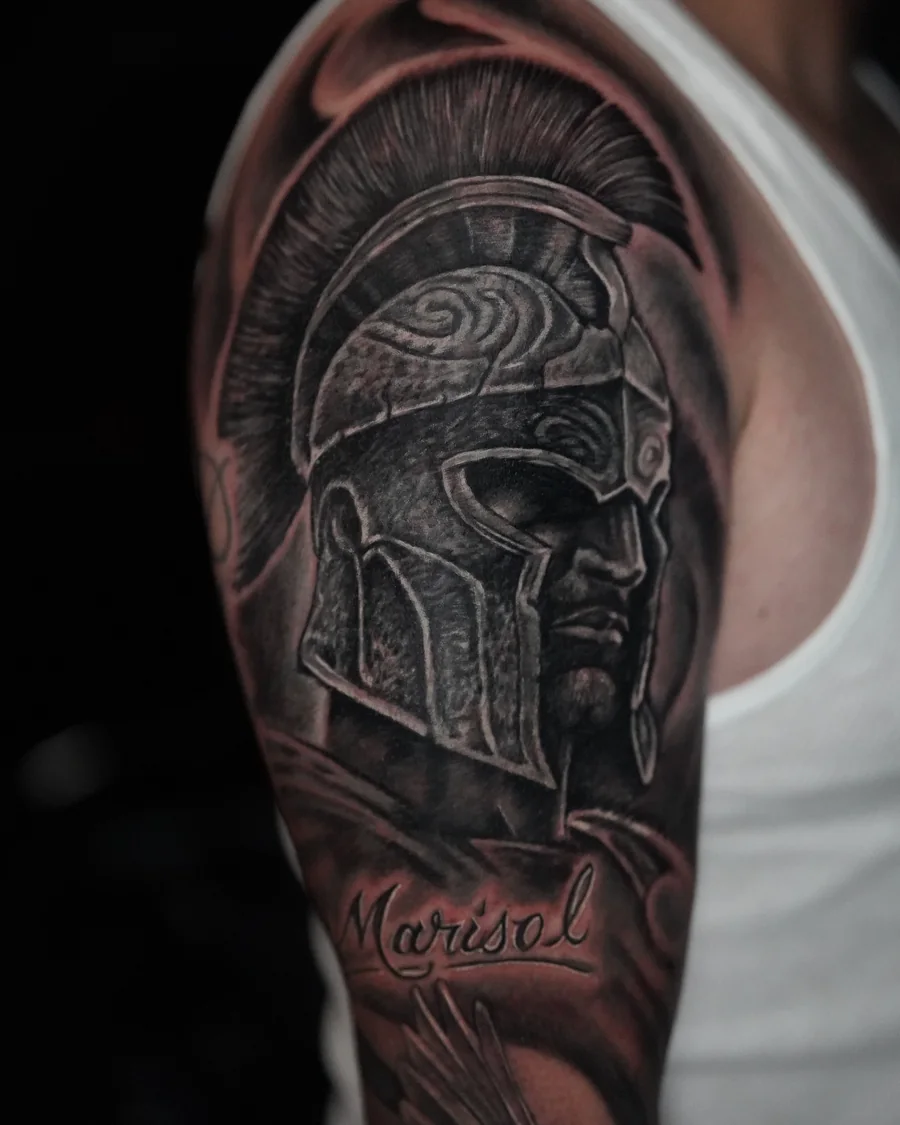 A black-and-gray upper-arm tattoo of a crested warrior helmet with engraved armor and a ribbon below.