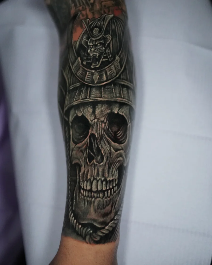 A realistic skull in a samurai helmet tattooed along the forearm, with a round crest above the brow.