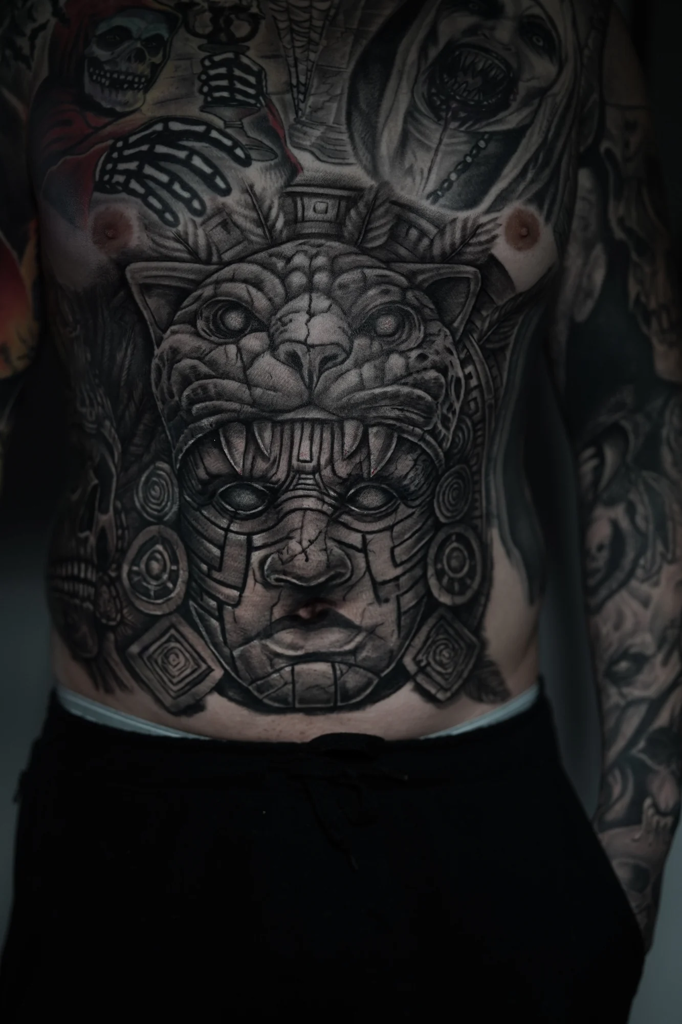 A large black-and-gray torso composition with a skull in a feathered headdress and a sculptural face framed by an animal headdress.