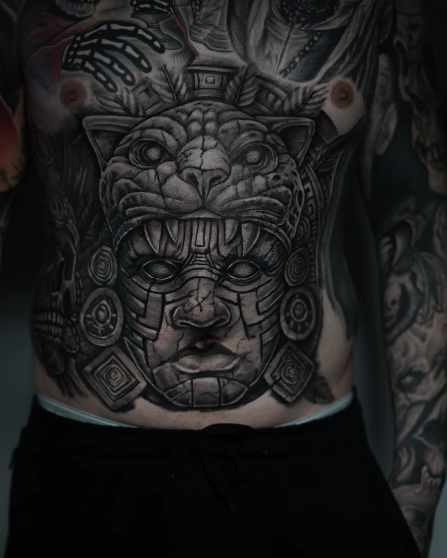 A large black-and-gray torso composition with a skull in a feathered headdress and a sculptural face framed by an animal headdress.