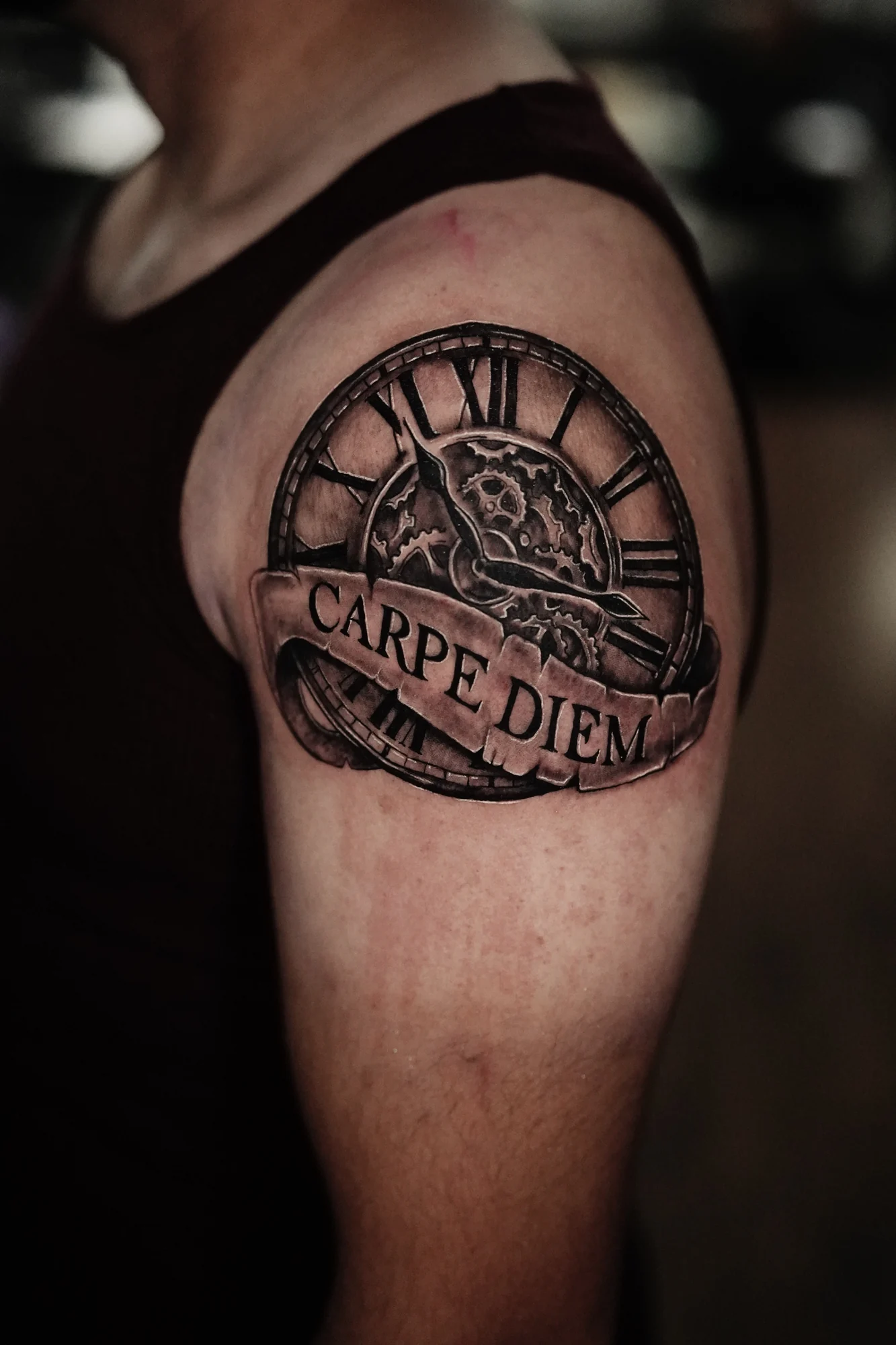 A circular clock tattoo with Roman numerals and a ribbon reading Carpe Diem on the upper arm.