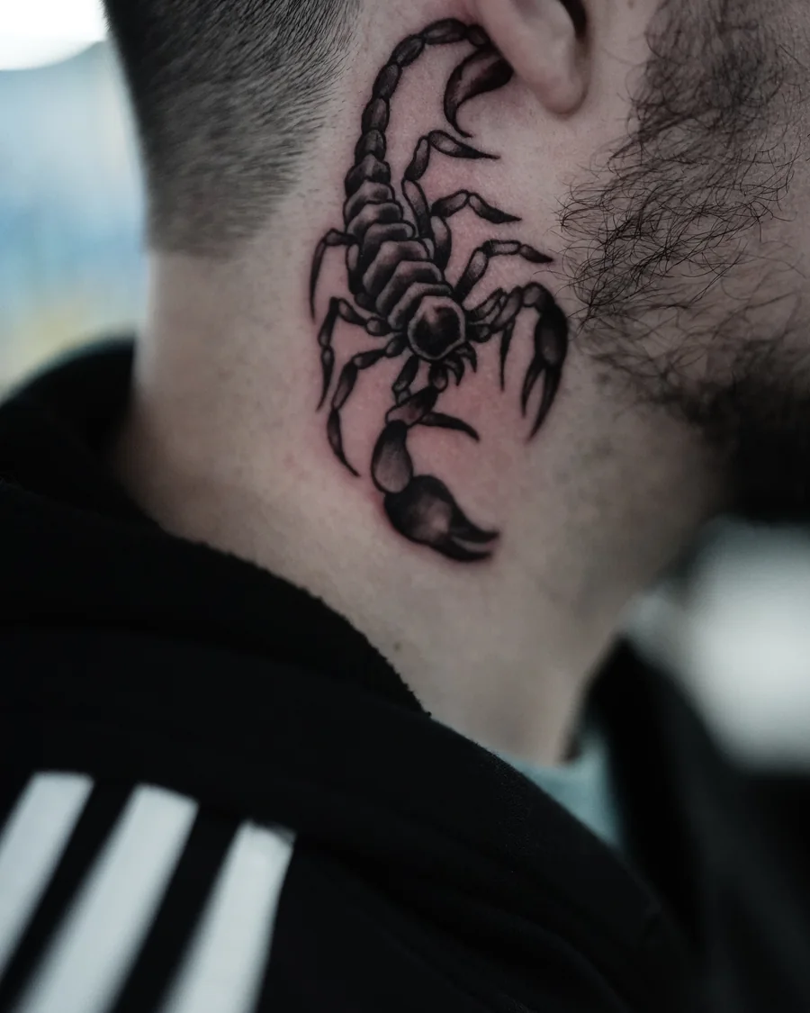 A small black scorpion tattoo behind the ear with its curved tail raised above its body.