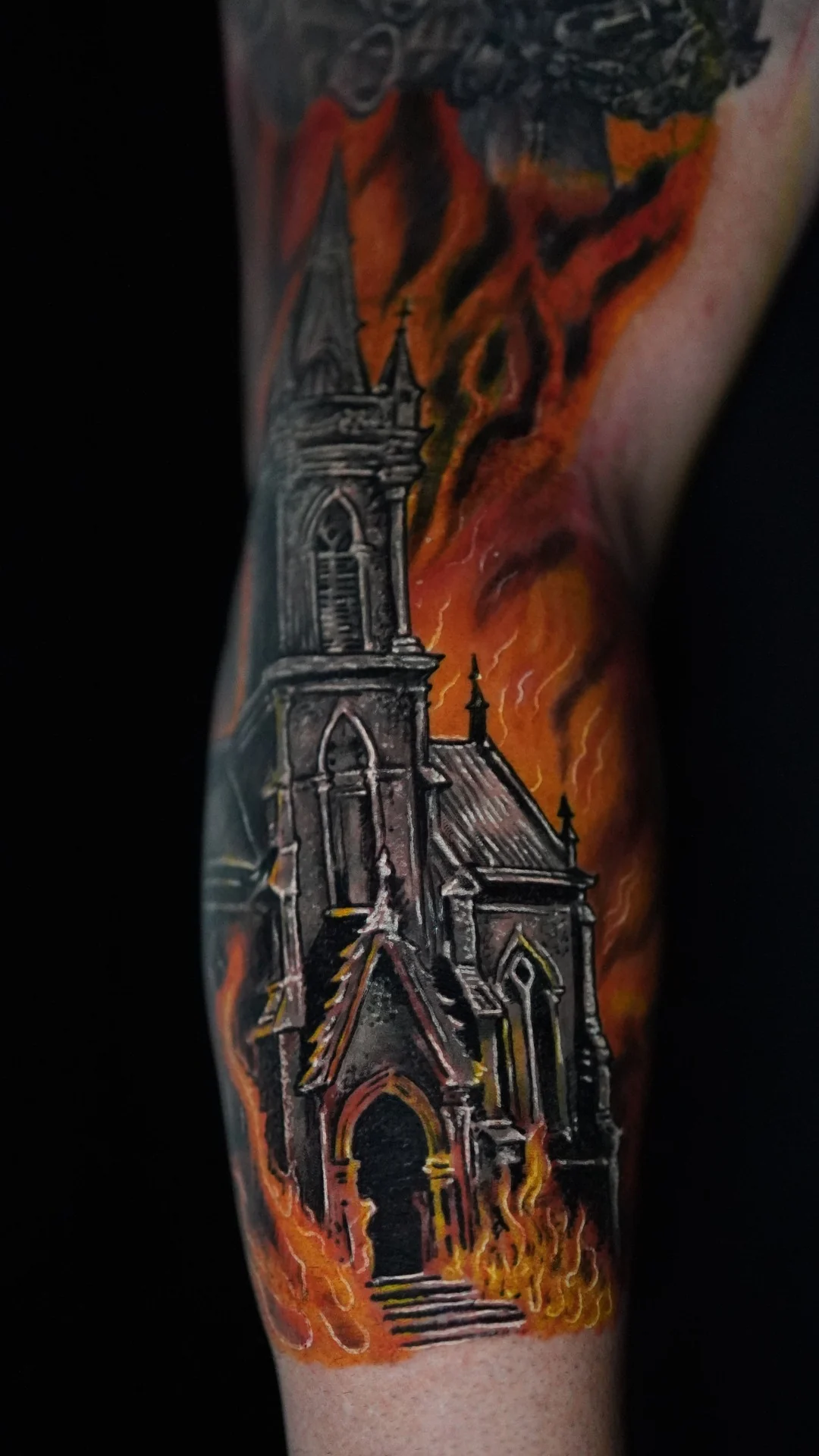 A tall church with a pointed steeple surrounded by orange flames in a vertical color tattoo.