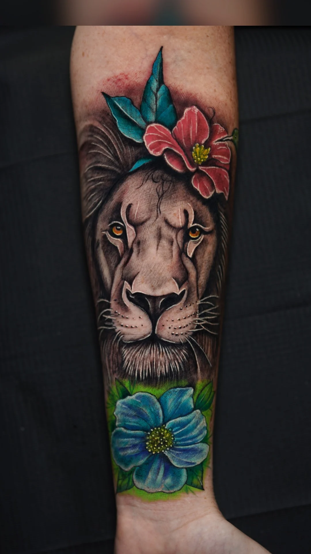 A lion portrait framed by red and blue flowers and green leaves in a vertical color tattoo.