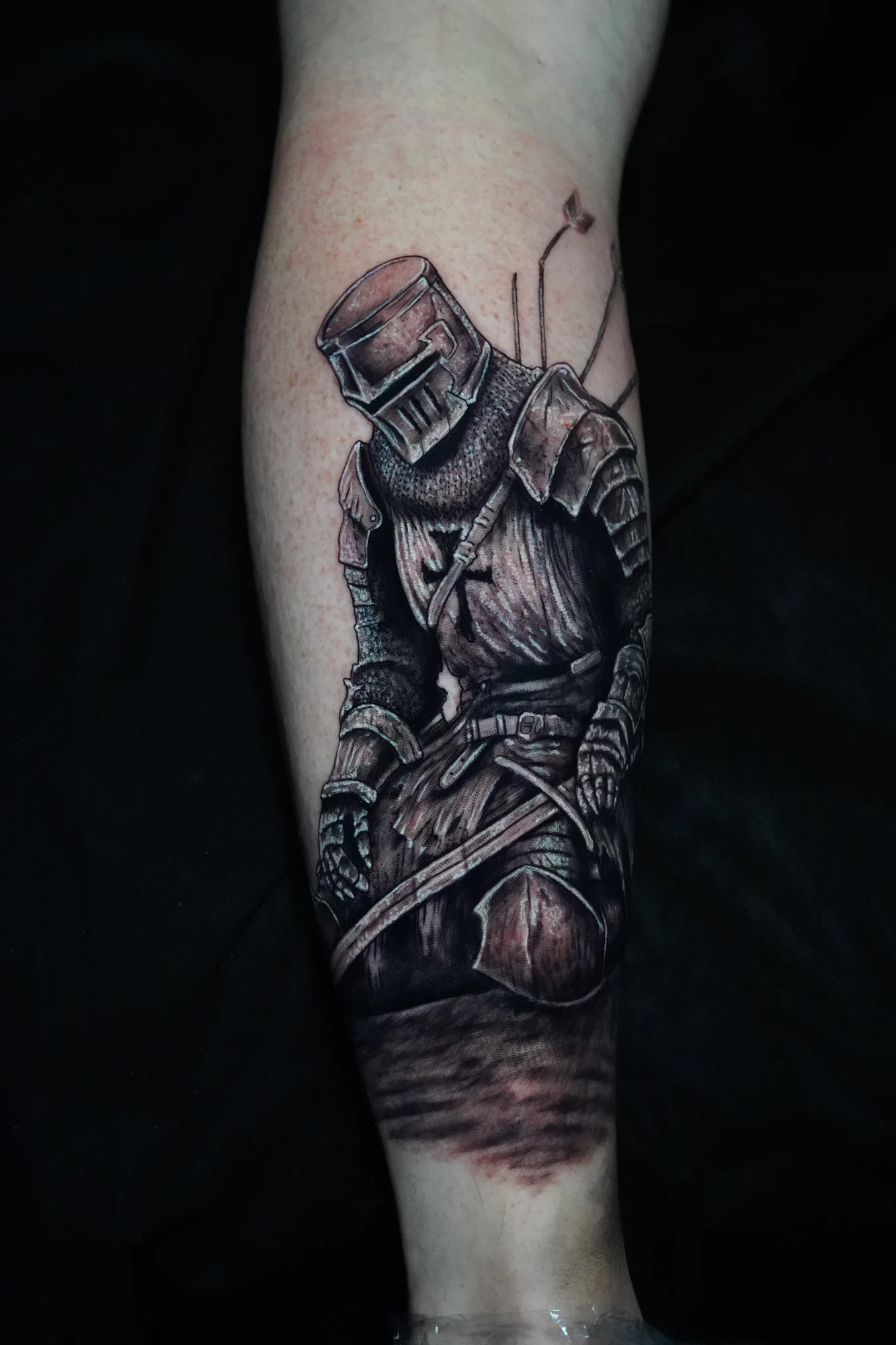 A kneeling armored knight with a closed helmet and sword, tattooed in black and gray on the forearm.
