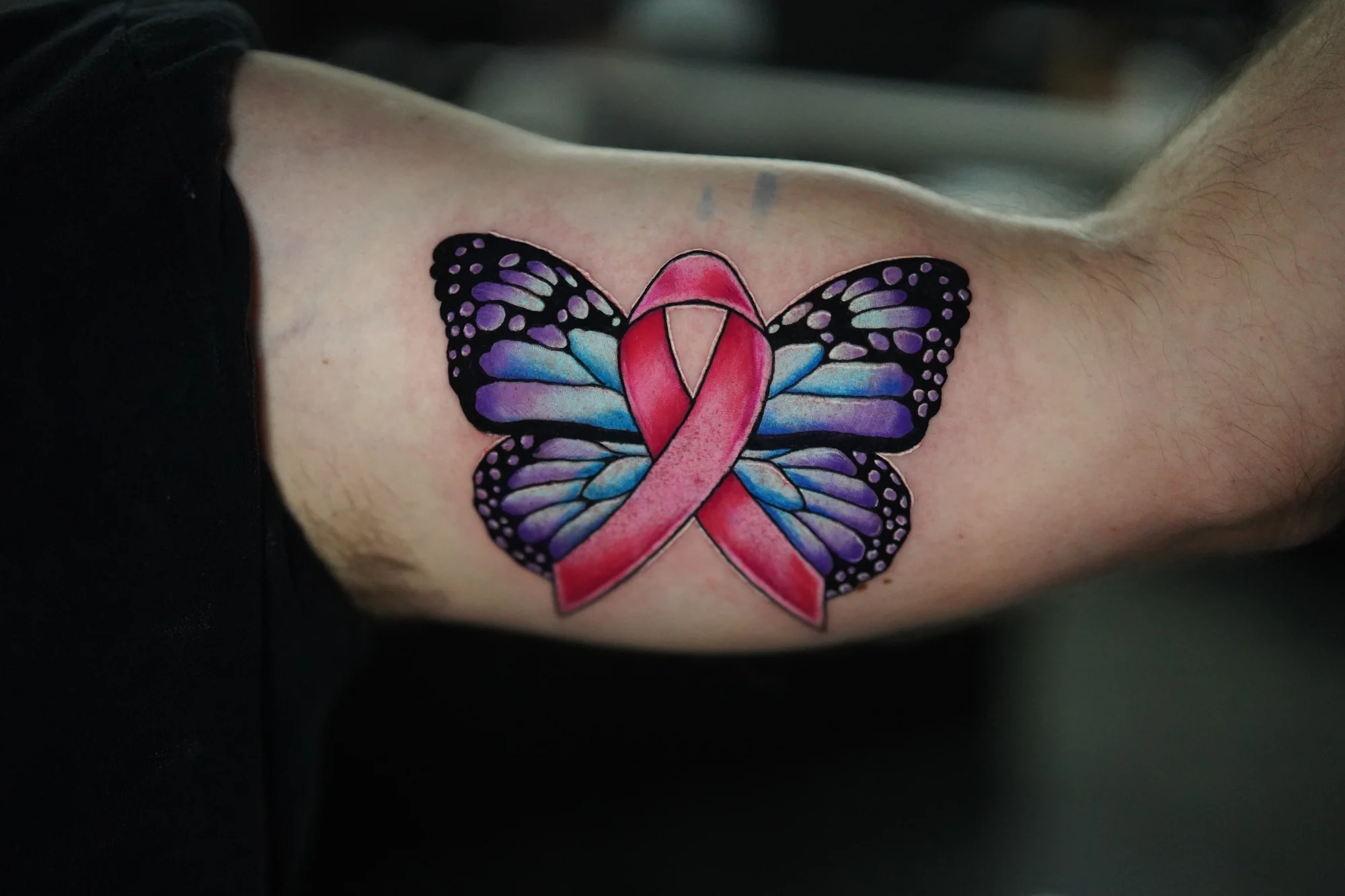 A butterfly with purple and blue wings surrounding a pink ribbon in a color tattoo.