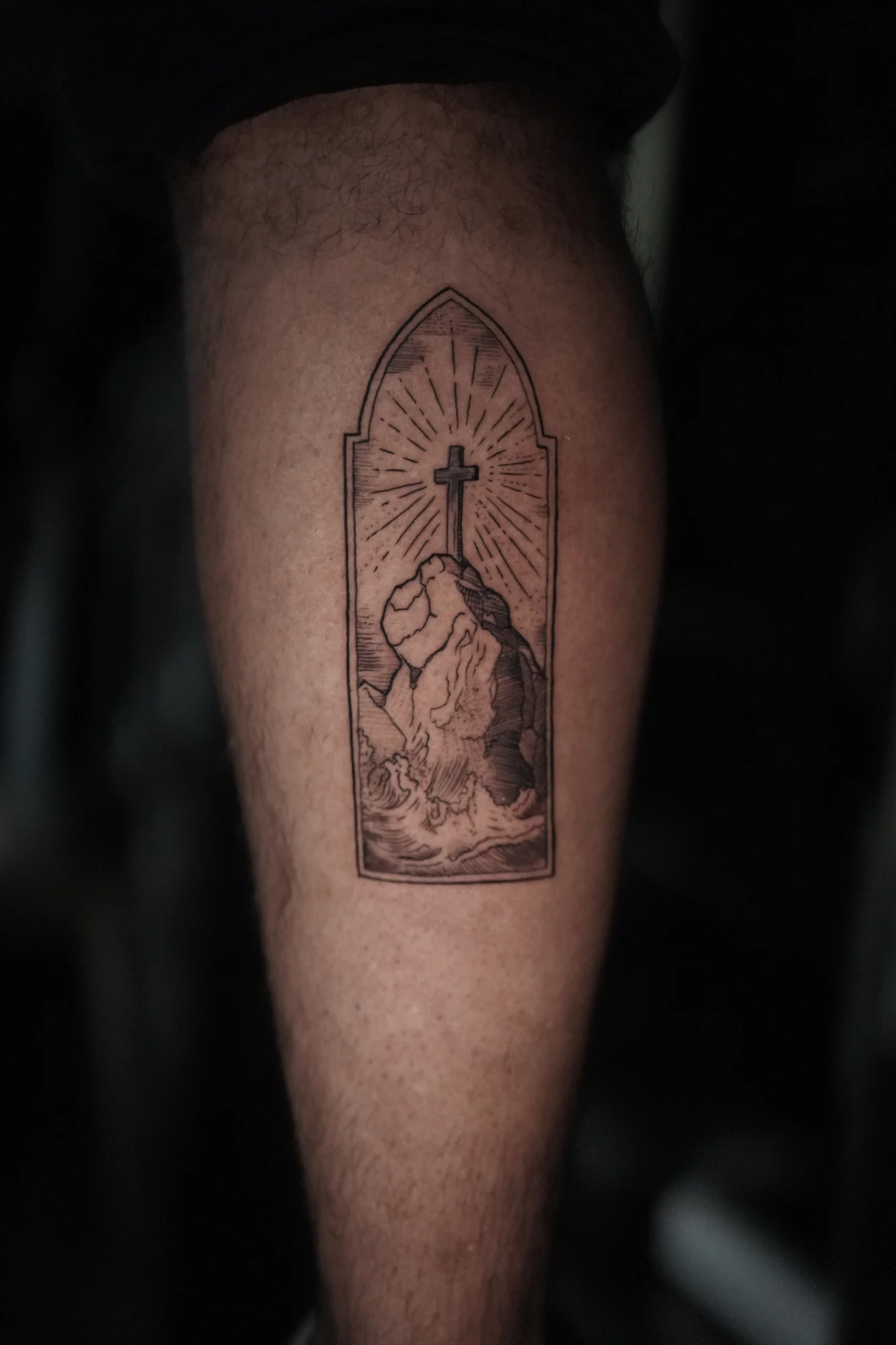 A pointed window-shaped tattoo enclosing clasped hands, a cross and rays of light in fine black linework.