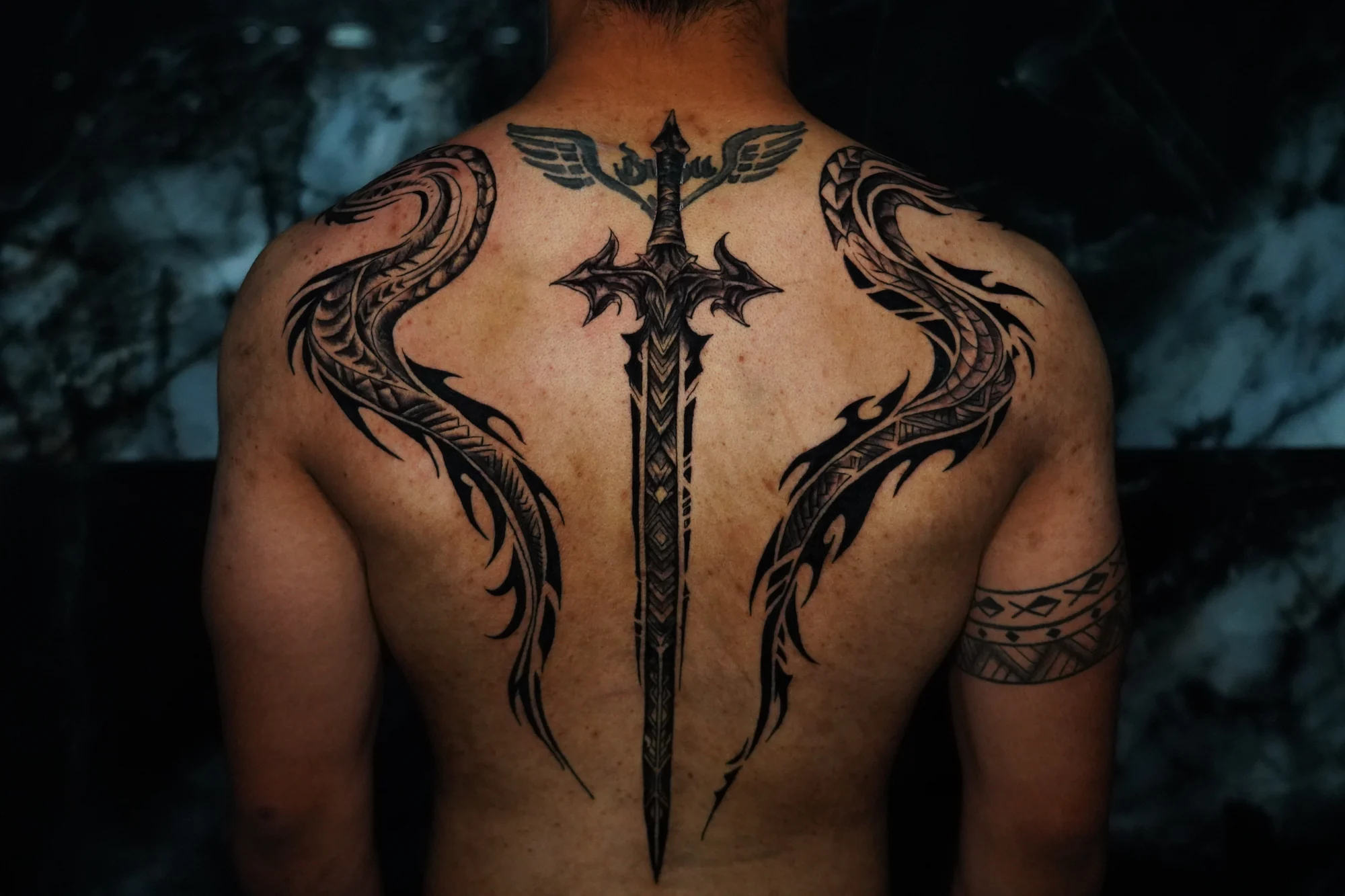Blackwork dragon tattoos extending from the chest over both shoulders to the back, with their bodies curving beside a central sword tattoo.