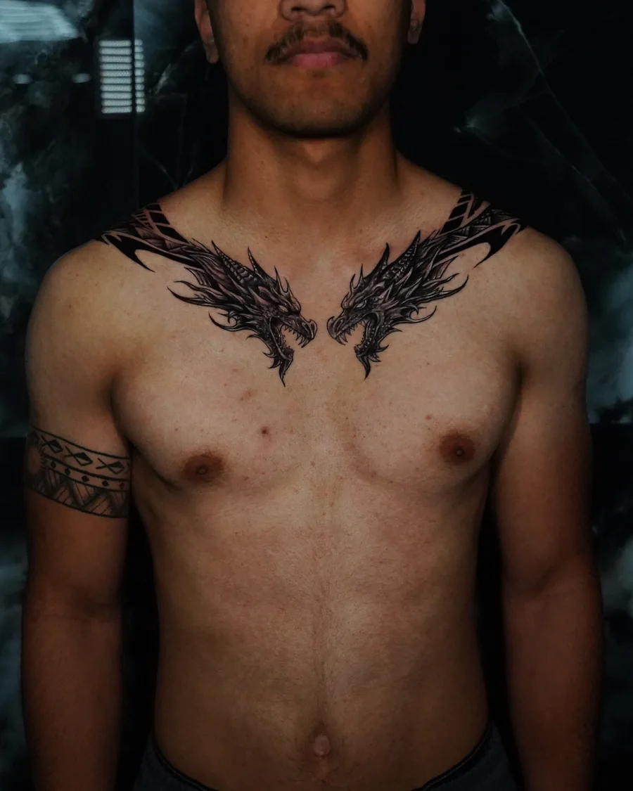 Front view of two blackwork dragon heads facing each other on the upper chest, continuing over the shoulders to the back, by William Martz.