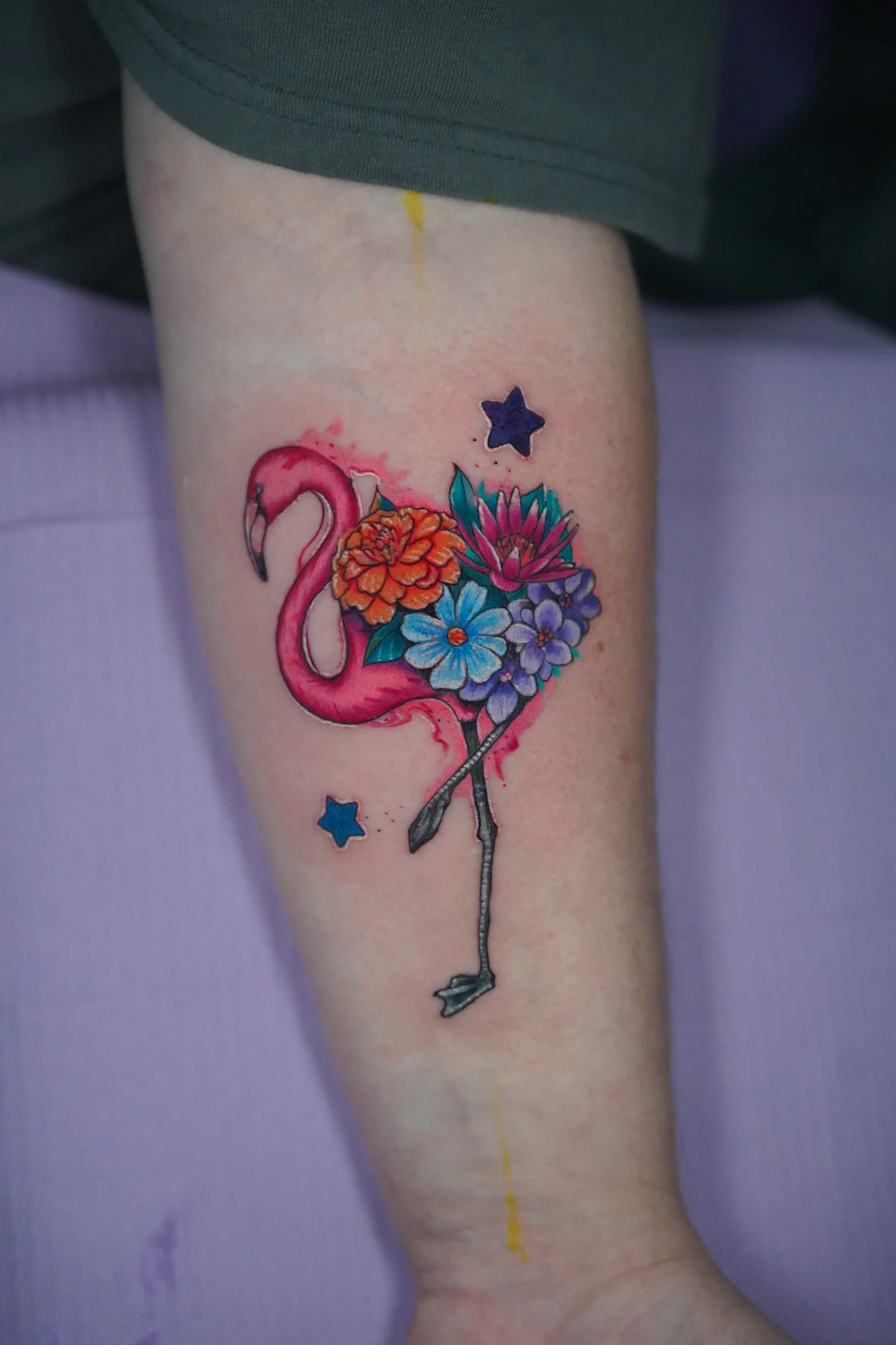 A pink flamingo tattoo with a cluster of colorful flowers and small star shapes around the bird.