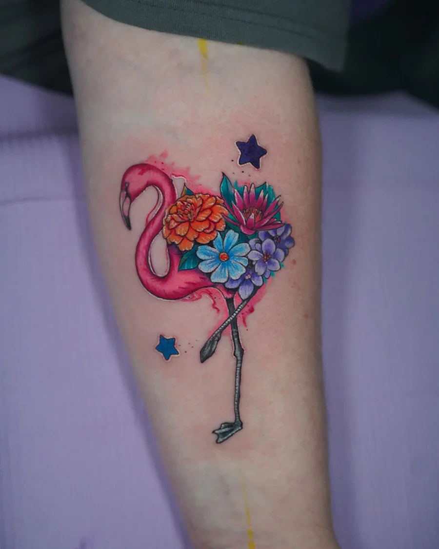 A pink flamingo tattoo with a cluster of colorful flowers and small star shapes around the bird.