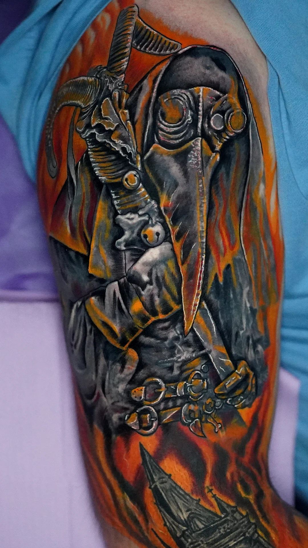 A winged skeletal figure wearing a gas mask, surrounded by orange flames in a color upper-arm tattoo.