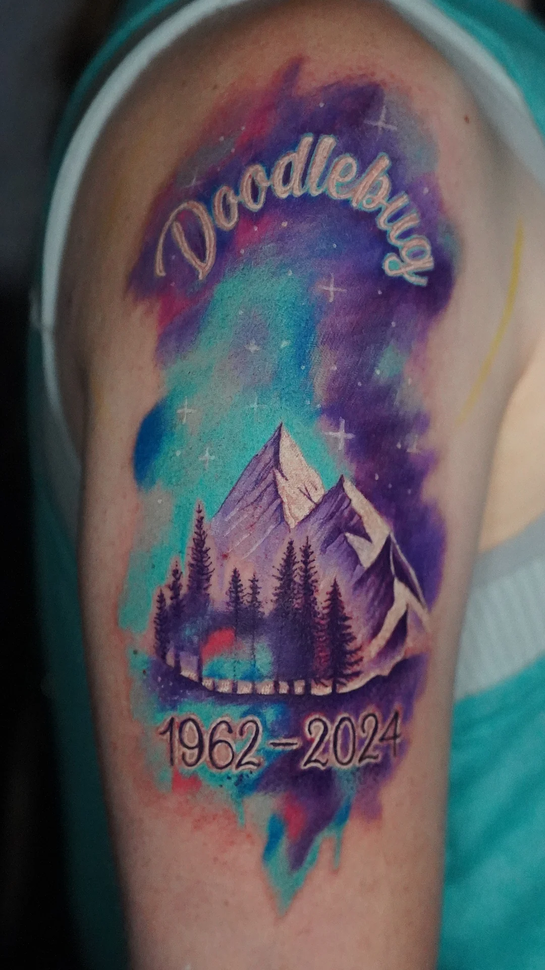 A mountain and forest tattoo under a purple, blue and green sky, with the word Doodlebug and dates framing the scene.