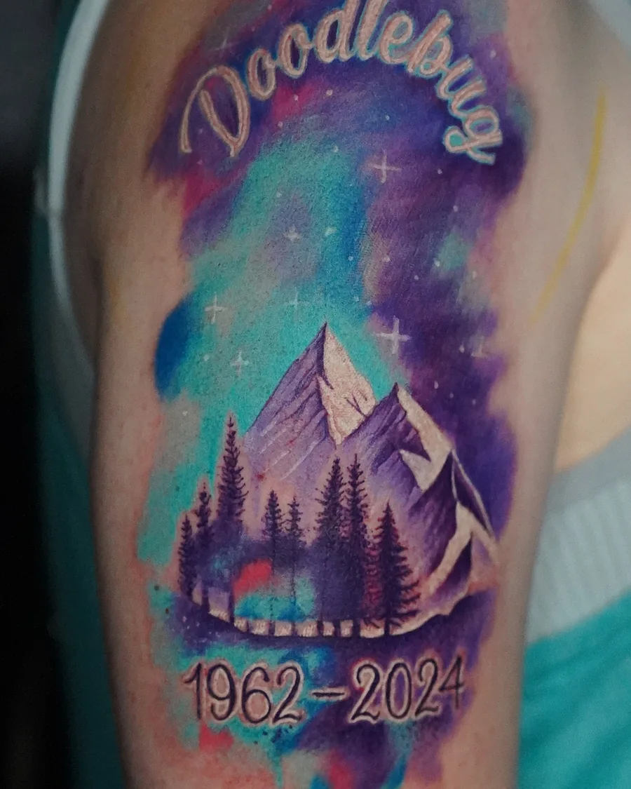 A mountain and forest tattoo under a purple, blue and green sky, with the word Doodlebug and dates framing the scene.
