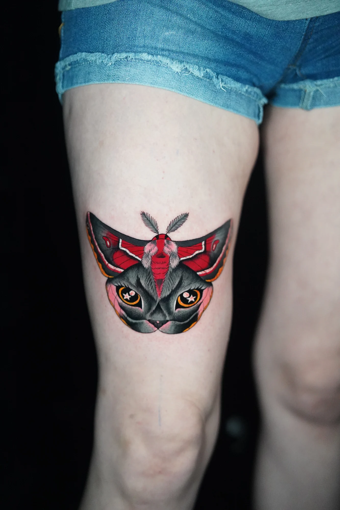 A symmetrical moth tattoo with red and black wings framing a dark cat face with bright eyes.