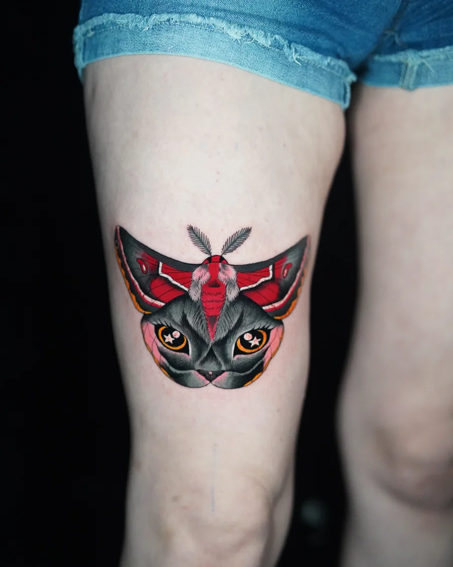 A symmetrical moth tattoo with red and black wings framing a dark cat face with bright eyes.