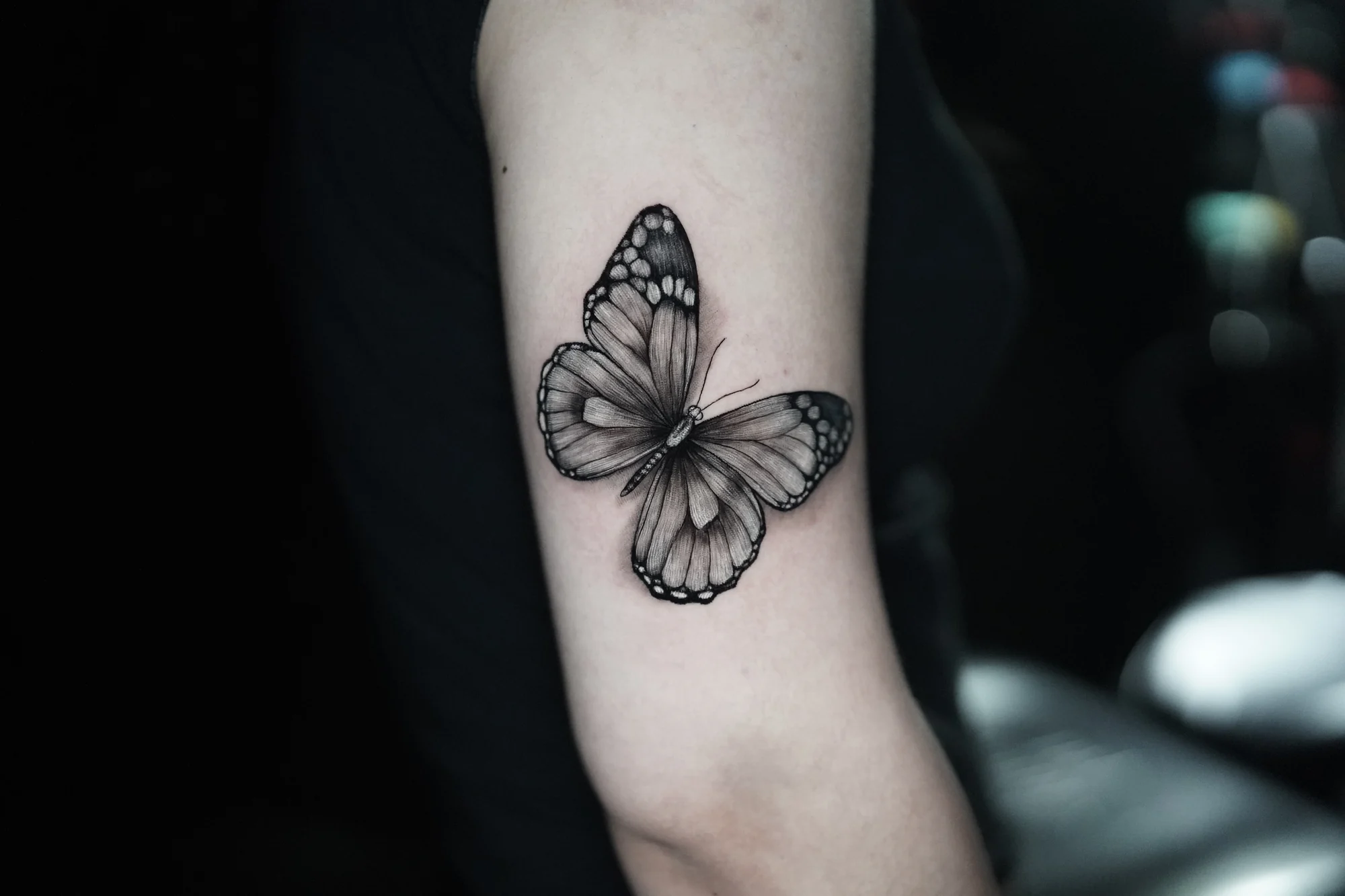 A black-and-gray butterfly tattoo with open wings, pale spots and fine wing veins on the arm.