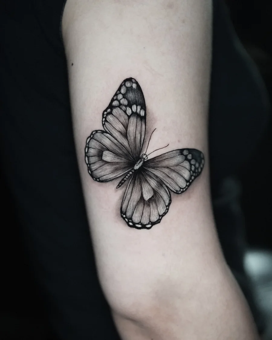 A black-and-gray butterfly tattoo with open wings, pale spots and fine wing veins on the arm.