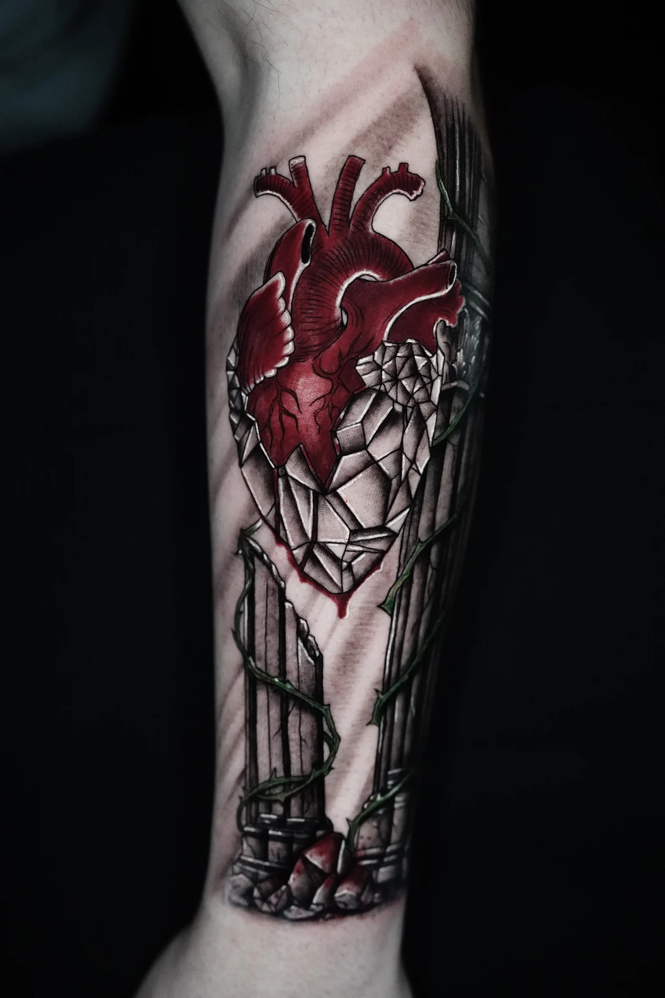A red anatomical heart above a pointed stained-glass-inspired window in a vertical forearm tattoo.