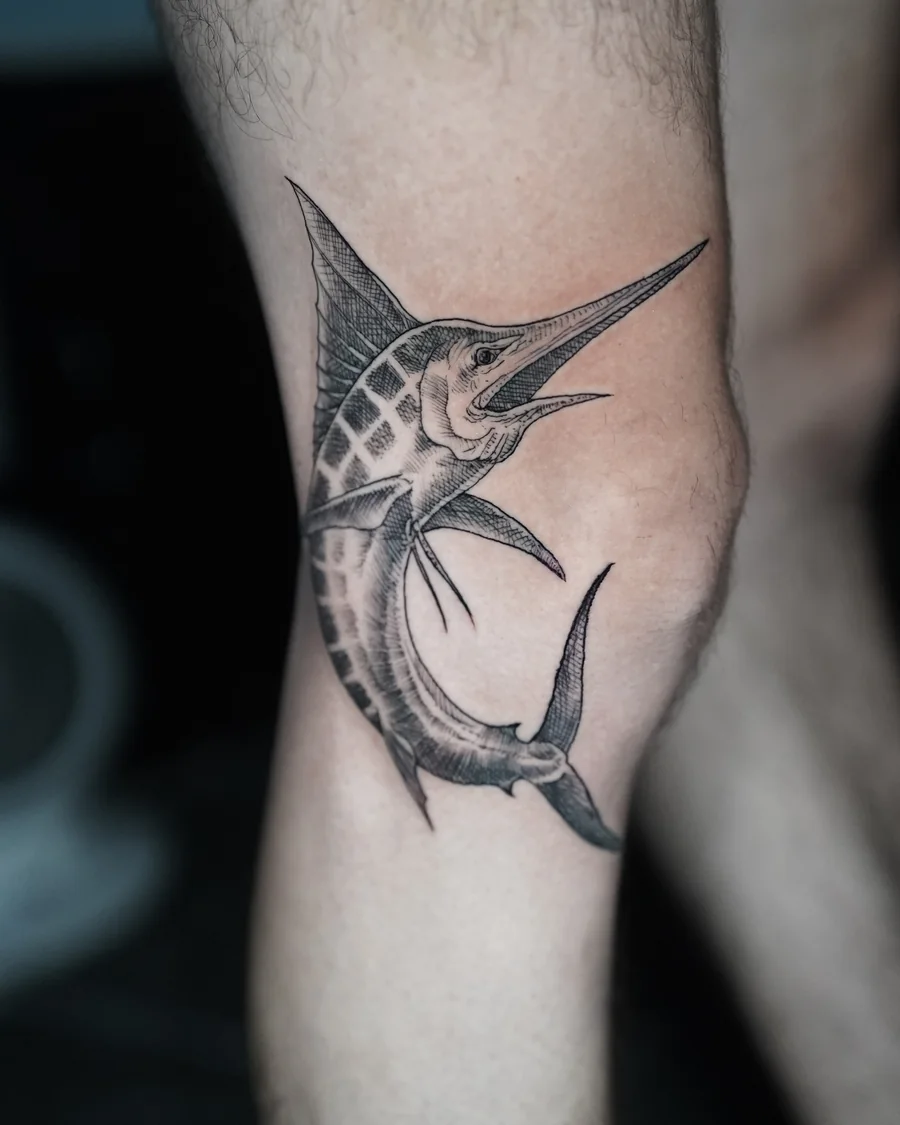 A curved marlin tattoo with a long pointed bill and a patterned back, rendered in black and gray on the arm.