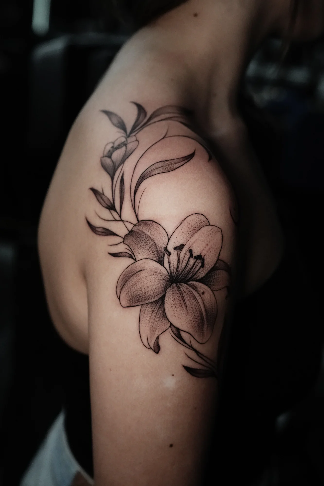 Large fine-line lilies and flowing leaves extending over the shoulder and toward the collarbone.