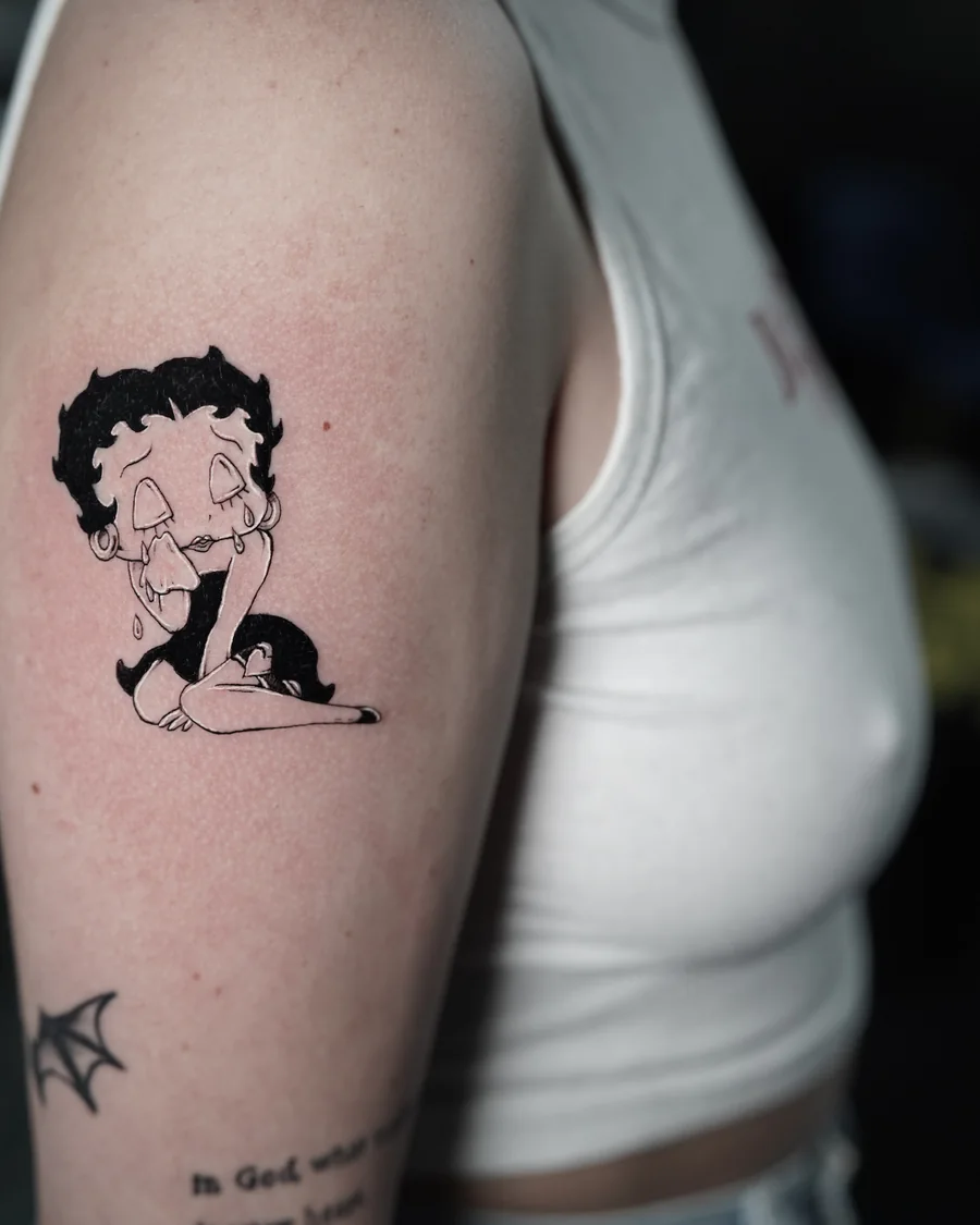 A small Betty Boop character tattoo in black linework and shading on the upper arm.