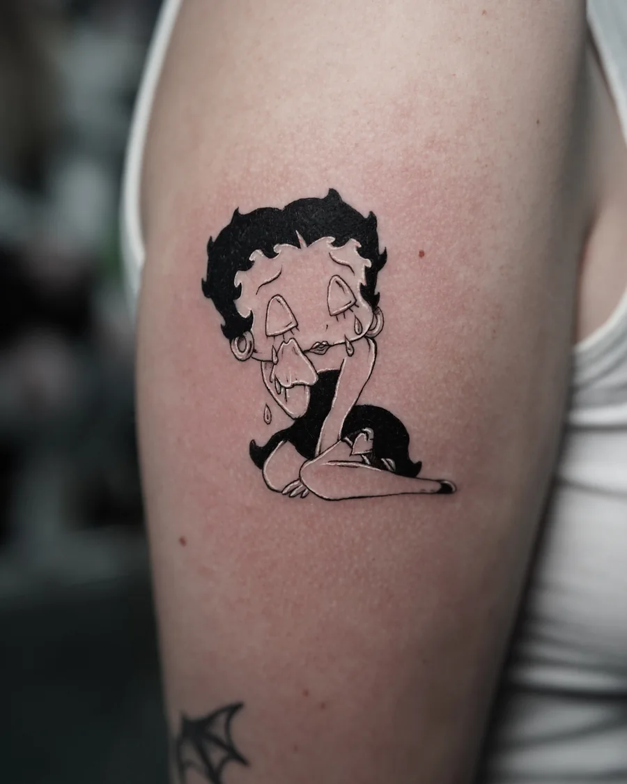 Alternate view. A small Betty Boop character tattoo in black linework and shading on the upper arm.