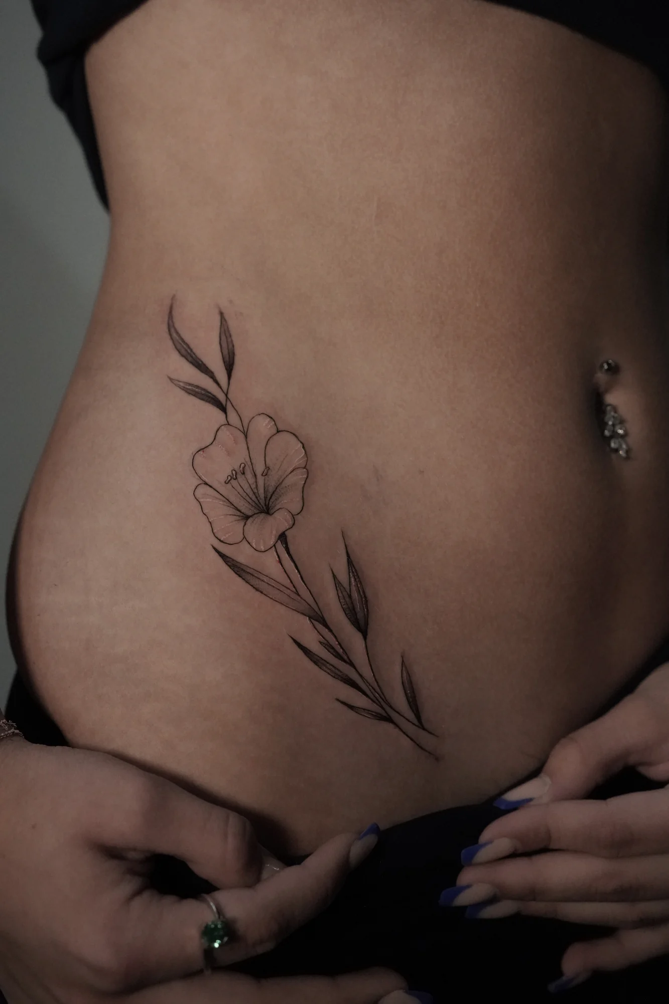 A single fine-line flower with long curved leaves placed along the side of the lower abdomen.