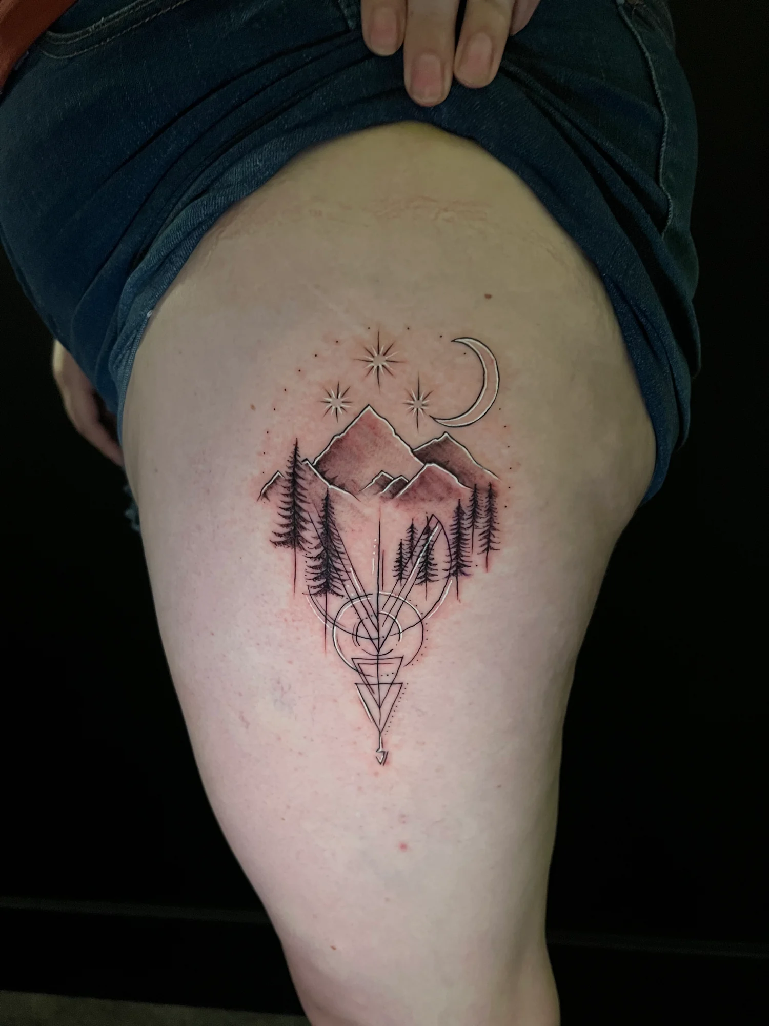 A geometric mountain and pine-tree tattoo with a crescent moon, small stars and pointed linework on the thigh.