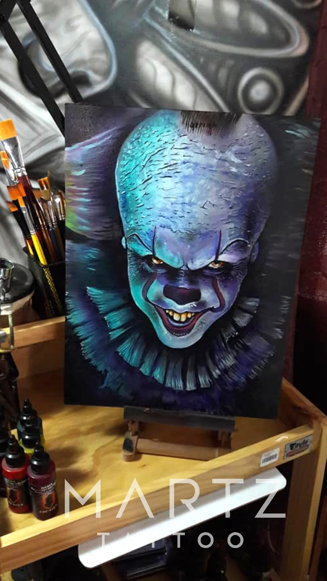 A painted Pennywise clown portrait with a pale blue forehead, yellow eyes and dark violet shadows on a canvas easel.