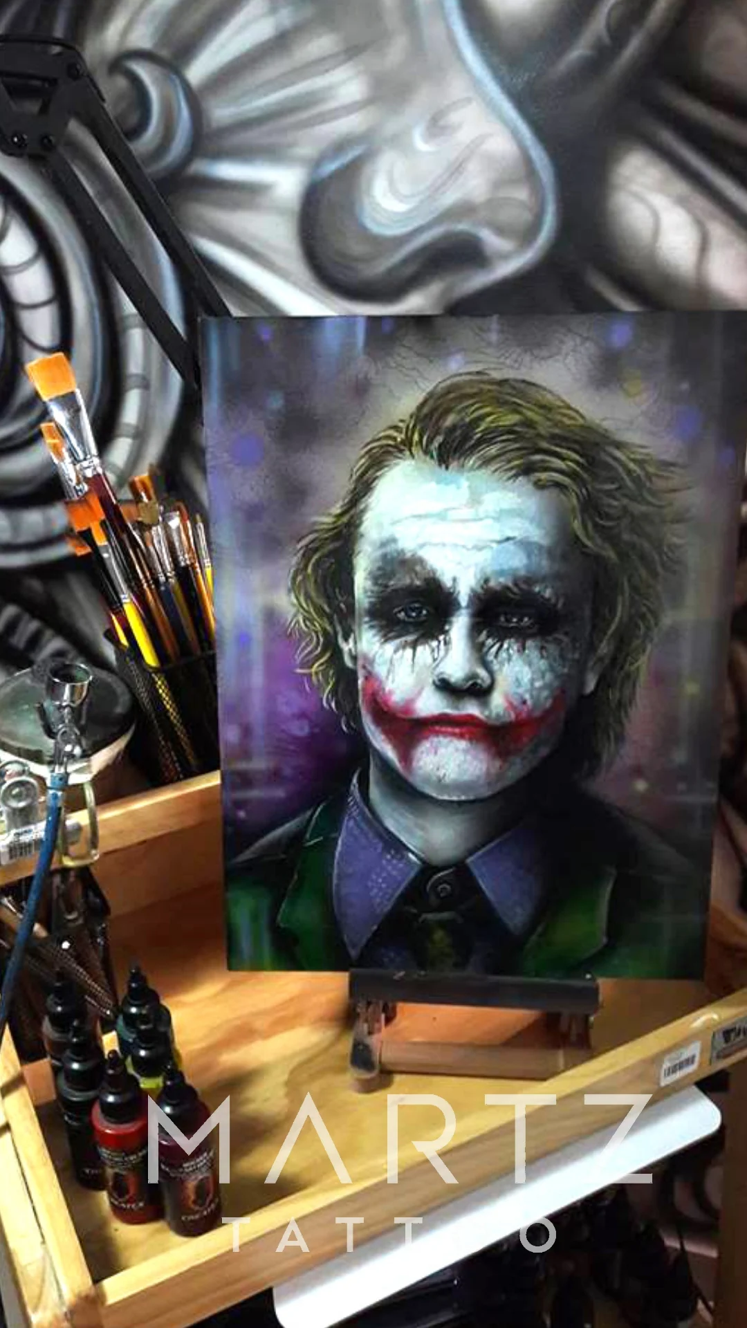 A painted Joker portrait with a white face, dark eyes, green hair and a red smile on a canvas easel.